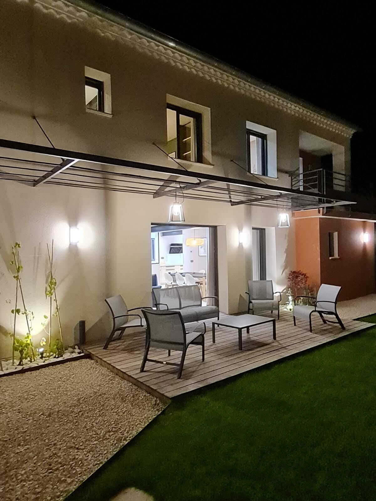 Un patio al aire libre con un sofá, sillas y una mesa, rodeado por una terraza de madera y un pequeño jardín.