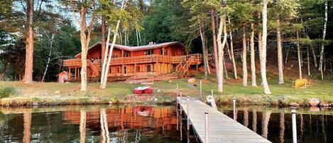 A wooden cabin with a dock extending into a calm lake, surrounded by trees.