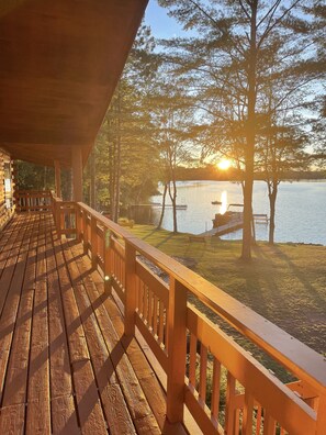 A wooden deck with a railing overlooking a lake, surrounded by trees and a dock with a boat.