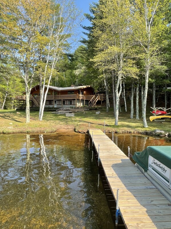 A wooden cabin with a deck and stairs leading to a porch, surrounded by trees and a calm body of water.