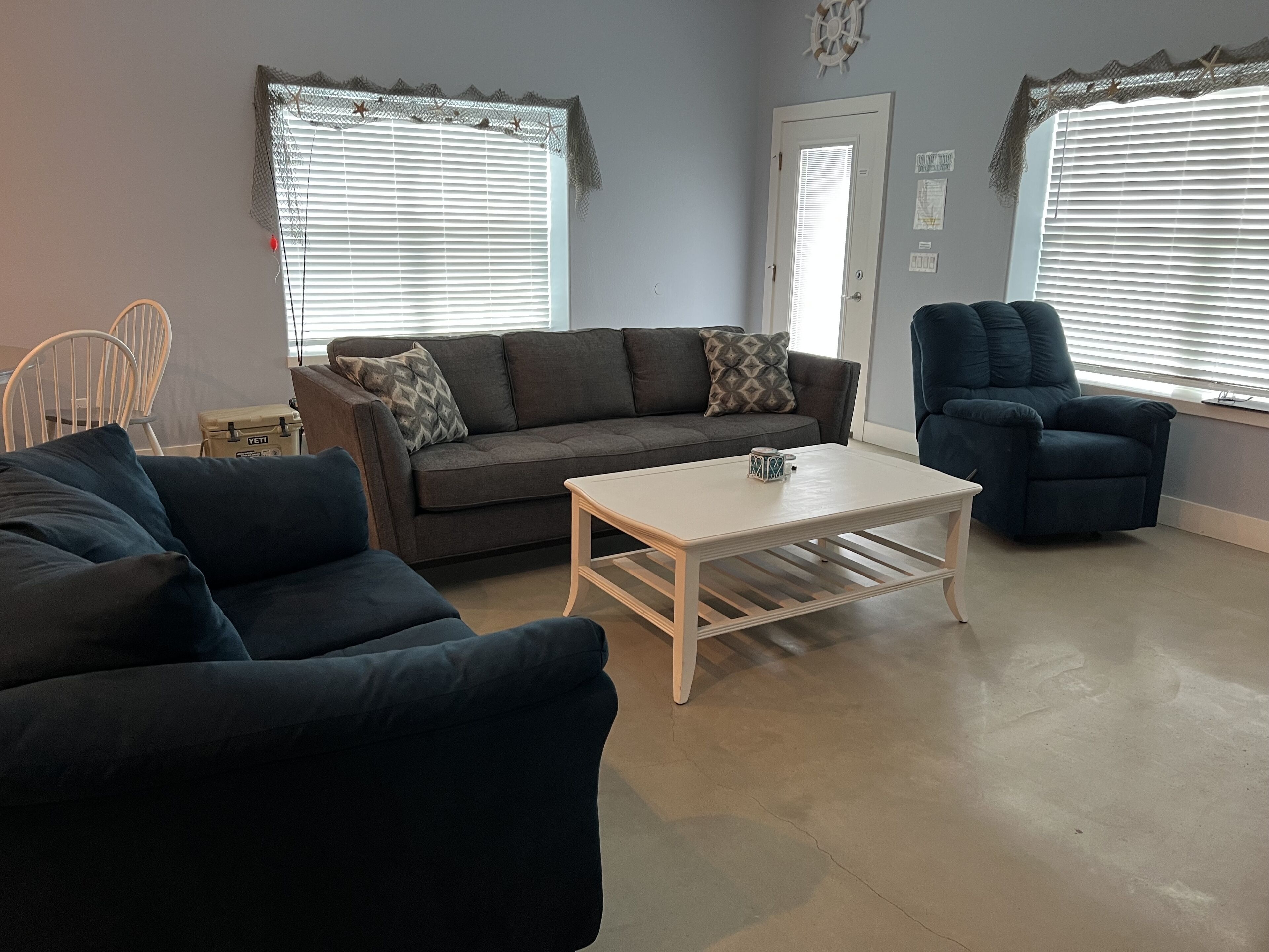 Un salon avec un canapé bleu foncé, une table basse blanche et un fauteuil bleu foncé. On y trouve une porte avec une fenêtre et une horloge murale.