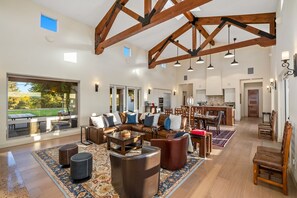 Amplia sala de estar con techo alto, vigas de madera, un ventanal con vistas al exterior, un comedor y una cocina.