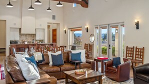 Un vaste espace de vie comprenant un canapé en cuir marron, une table basse en bois et un coin repas avec des chaises en bois. On aperçoit une cuisine au fond, et de grandes fenêtres offrent une vue sur l’extérieur.