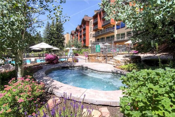 Un jacuzzi rodeado de un jardín con flores y árboles, con un edificio al fondo.