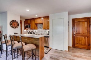 Una cocina con armarios de madera, microondas, estufa y lavabo. Hay cuatro taburetes de bar y un reloj en la pared.
