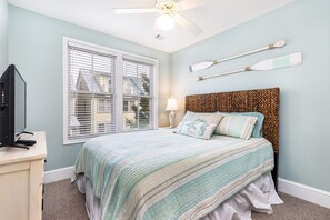 Une chambre à coucher avec un lit, un téléviseur posé sur un meuble, une fenêtre avec des stores et un ventilateur de plafond.