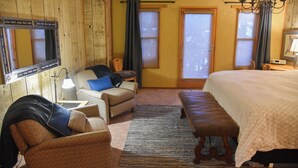 Una habitación con cama, una silla, un banco, un espejo y dos ventanas con cortinas.