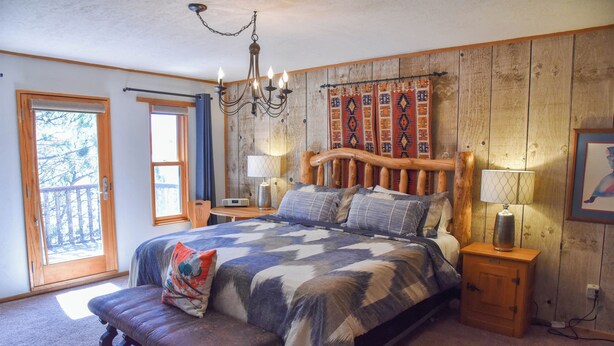 Un dormitorio con cabecera de madera, una cama grande con colcha estampada, un banco al pie de la cama y una mesita de noche con lámpara.