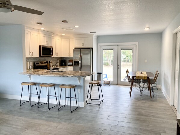 A modern kitchen with white cabinets, a stainless steel refrigerator, and a granite countertop. There are three stools at the island and a dining table with chairs.