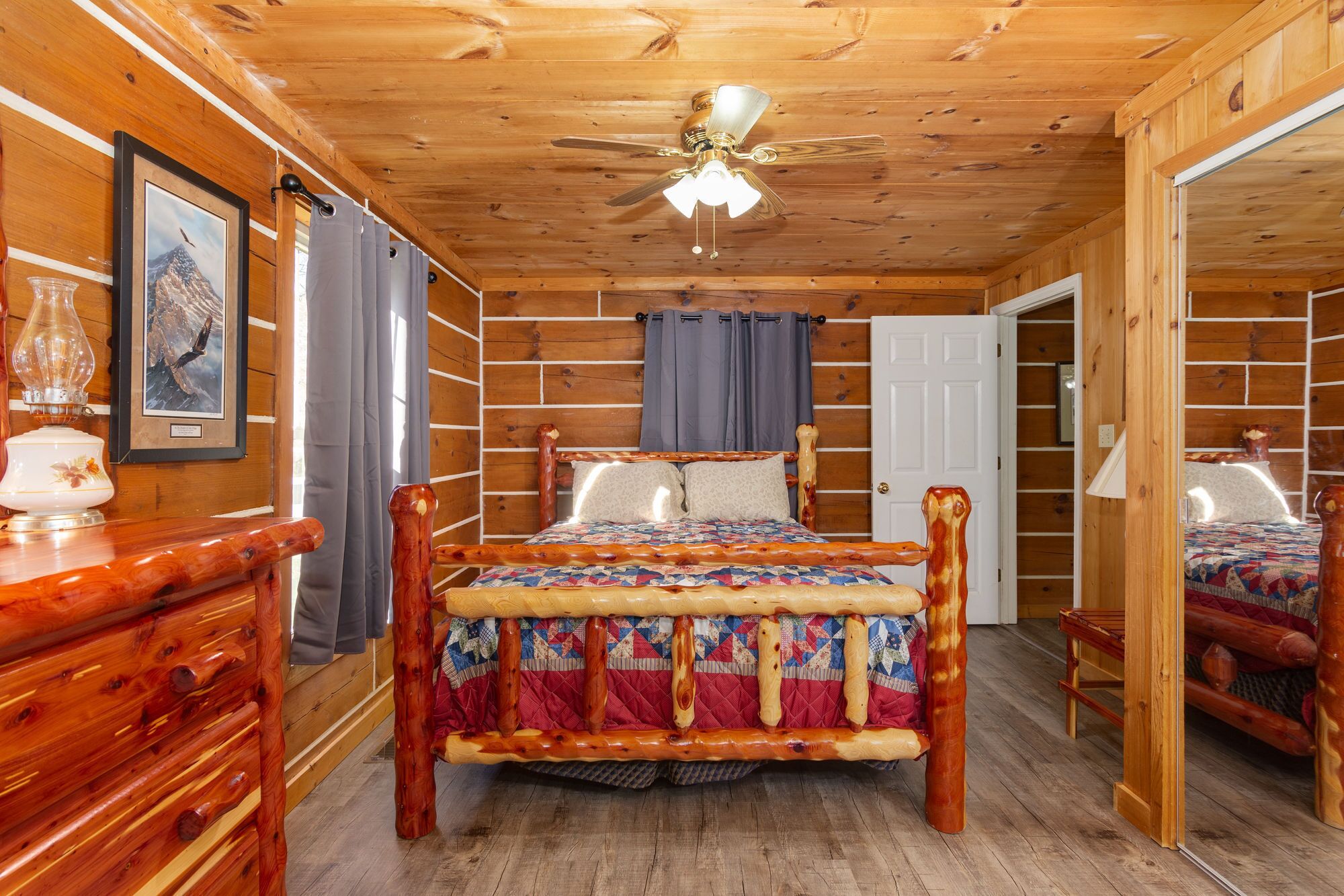 A wooden cabin bedroom with a bed, a ceiling fan, and a door leading to another room.
