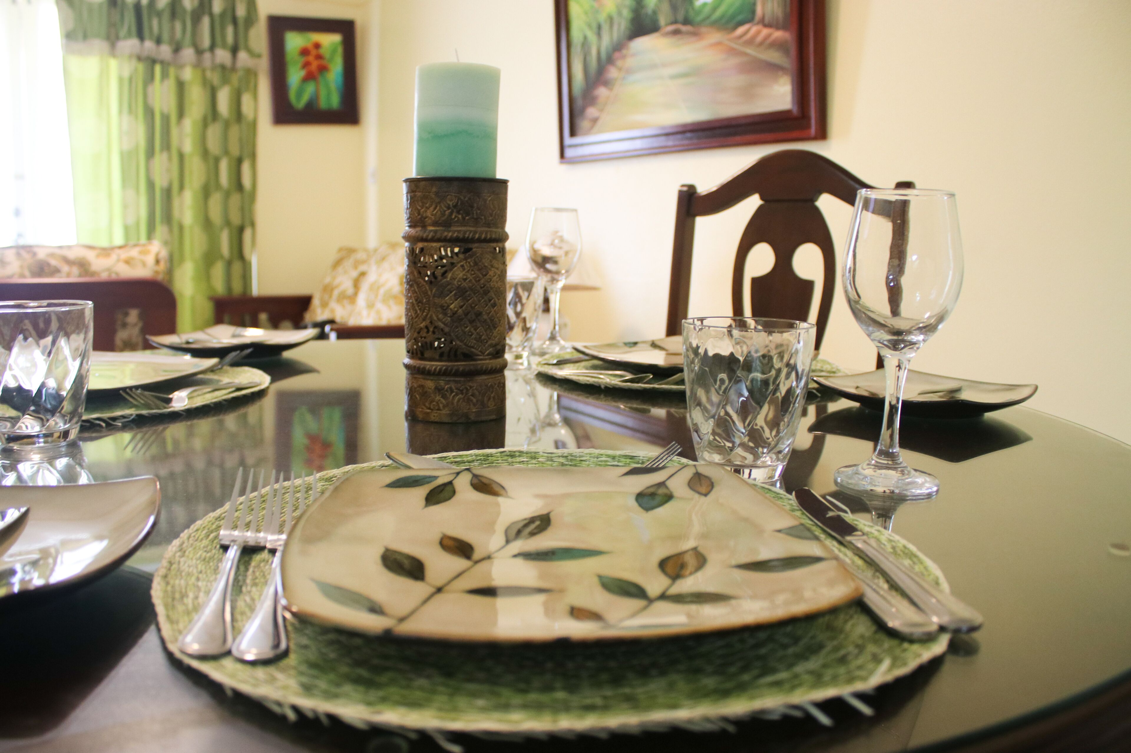 Una mesa de comedor dispuesta con platos, cubiertos y vasos, con una vela y un cuadro al fondo.