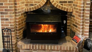 A brick fireplace with a clock above it, a fire burning inside, and a bag of firewood on the right.