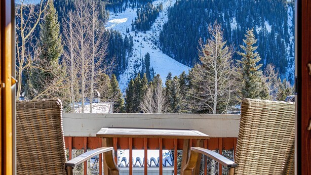Un balcon avec des fauteuils et une table en osier, offrant une vue sur un paysage montagneux enneigé, avec des arbres et une piste de ski.
