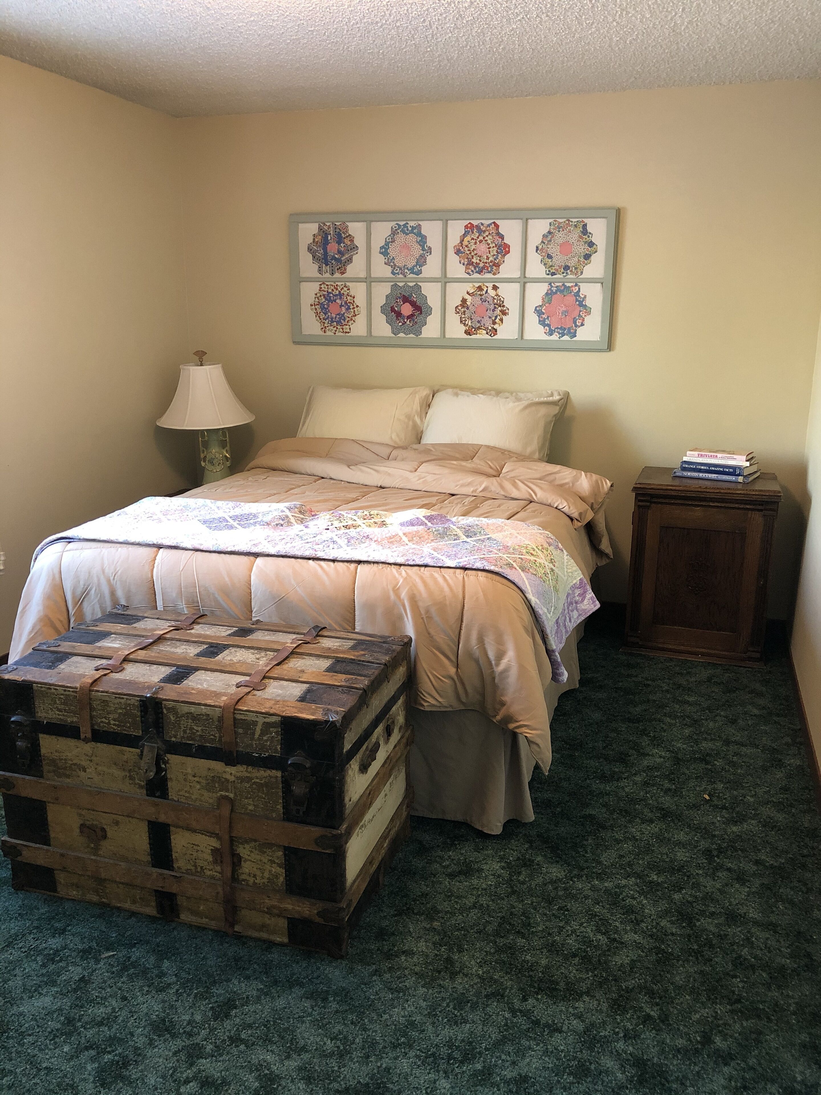 A bedroom with a bed, a quilt, a lamp, a chest of drawers, and a wall with framed artwork.
