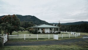 A single-story house with a green roof, surrounded by a white fence, set in a rural area with hills in the background.