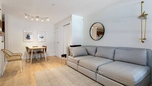 A modern living room with a grey sofa, a dining table with chairs, a small rug, and a wall-mounted mirror.