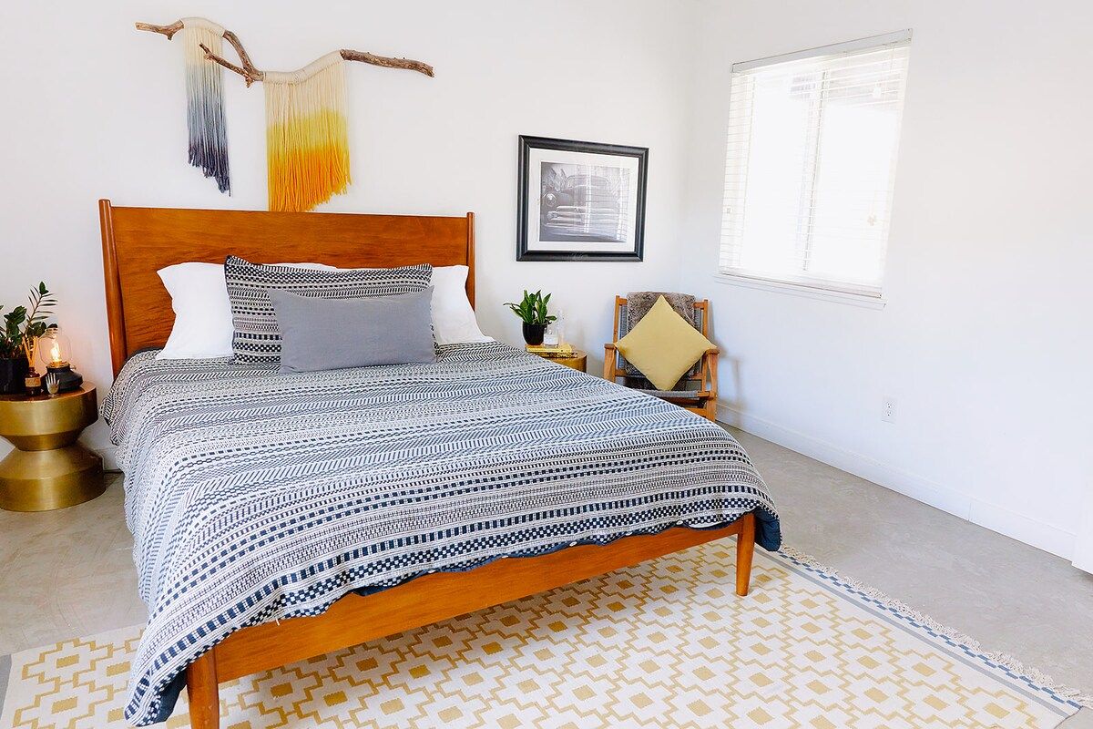 A bed with a patterned bedspread, a wooden headboard, a bedside table with a plant, and a window with blinds.