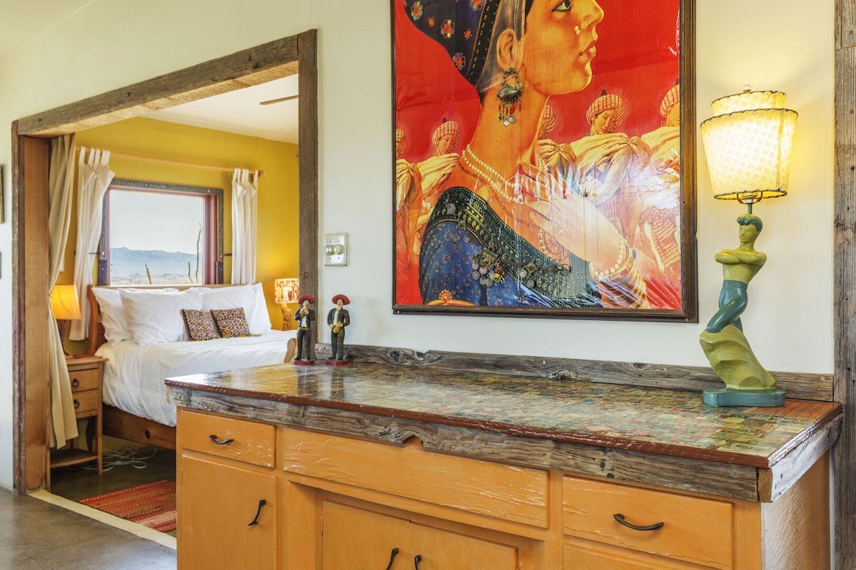 A hotel room with a bed, a wooden dresser, a painting, and a lamp.