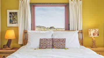 Ein Bett mit weißer Bettwäsche, zwei gemusterte Kissen, ein Holz-Kopfstück, ein Fenster mit Vorhängen, ein Nachttisch mit Lampe und ein gerahmtes Bild an der Wand.
