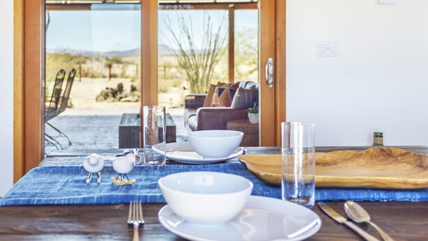 A dining table set with white dishes, a glass, and cutlery, with a view of a desert landscape through large glass doors. - 🌵MoonView Modern | Hot tub, BBQ & Private Pool🌵 (Joshua Tree)