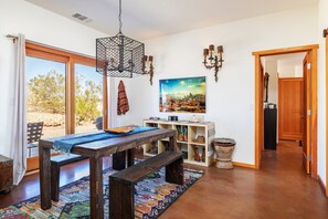 Una habitación con una mesa de madera, bancos, una lámpara colgante, un estante con adornos y una puerta que da a otra estancia.