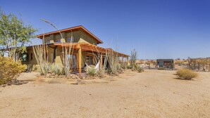 A modern house with a flat roof and a covered porch, surrounded by desert vegetation. - MoonView Modern | Hot tub, BBQ & Private Pool! (Joshua Tree)