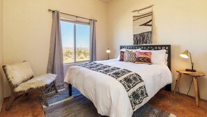 A bedroom with a large bed, a chair, a nightstand, a lamp, and a view of the desert through the window. - Horizon House | Hot Tub, Fire Pit & BBQ in JT (Joshua Tree)