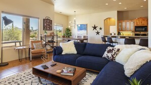 A modern living room with a blue sofa, a coffee table, and a dining area with a kitchen in the background. - 🌵 Horizon House | Hot Tub, Fire Pit, Games & BBQ | Adobe in JT 🌵 (Joshua Tree)