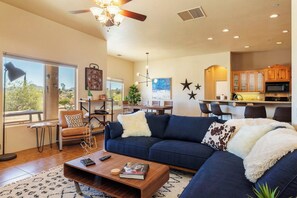A modern living room with a sectional sofa, a coffee table, and a dining area with a kitchen in the background. - Horizon House | Hot Tub, Fire Pit & BBQ in JT (Joshua Tree)