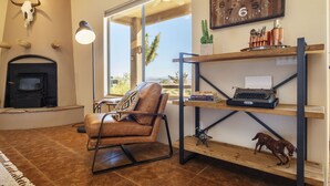 A cozy living room with a fireplace, a leather chair, a wooden shelf with books and a typewriter, and a large window with a view. - 🌵 Horizon House | Hot Tub, Fire Pit, Games & BBQ | Adobe in JT 🌵 (Joshua Tree)