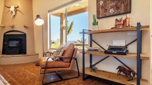 A living room with a fireplace, a leather chair, a bookshelf, and a large window with a view. - Horizon House | Hot Tub, Fire Pit & BBQ in JT (Joshua Tree)