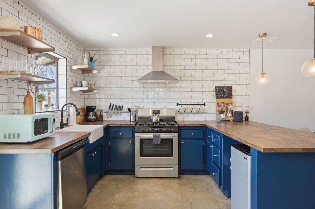 Una cocina con estufa de acero inoxidable, armarios azules, salpicadero de azulejos blancos y encimera de madera.