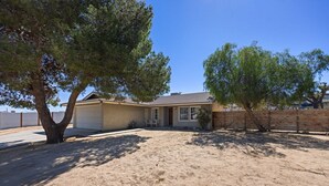 A single-story house with a garage, surrounded by a sandy yard and two trees. - Cosmic Rabbit - Family Friendly home w/Hot Tub (Joshua Tree)