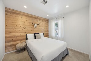 A bedroom with a wooden headboard, a large bed, a small stool, and a window with curtains. - Cosmic Rabbit - Family Friendly home w/Hot Tub (Joshua Tree)