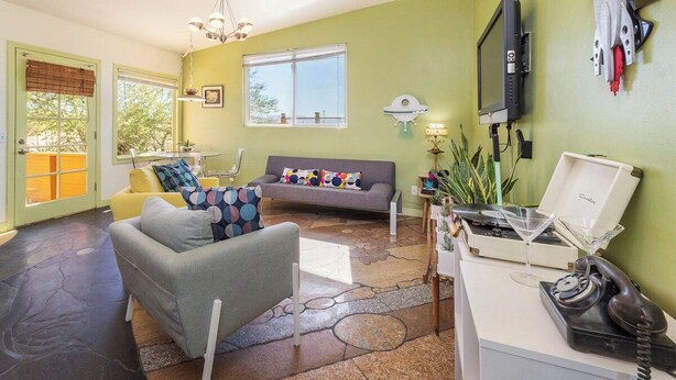 Una sala de estar con sofá, sillón y una mesa de centro. Hay un televisor montado en la pared, un teléfono y un objeto decorativo que se asemeja a una nave espacial.