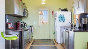 Cocina compacta con electrodomésticos de acero inoxidable, una puerta verde y una pequeña zona de estar con una silla verde.