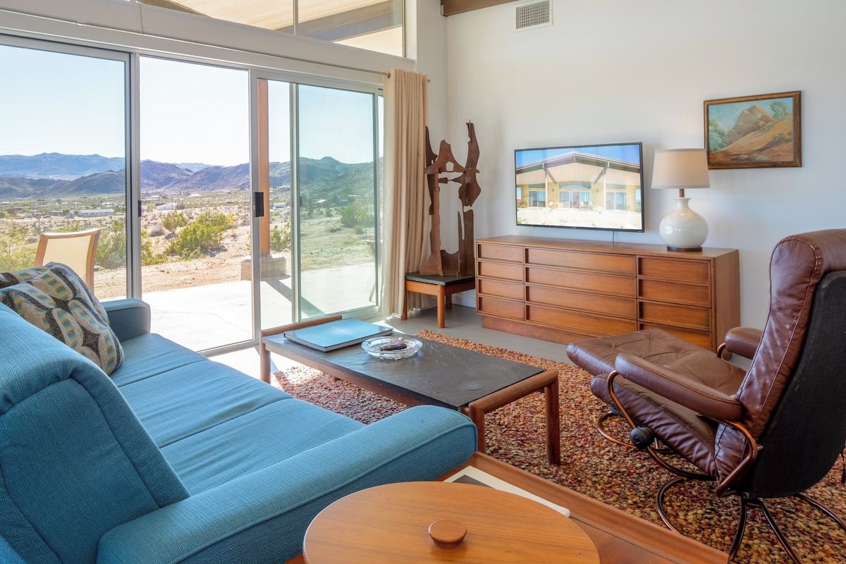 A living room with a blue sofa, a leather recliner, a wooden coffee table, and a view of the desert through large sliding glass doors.