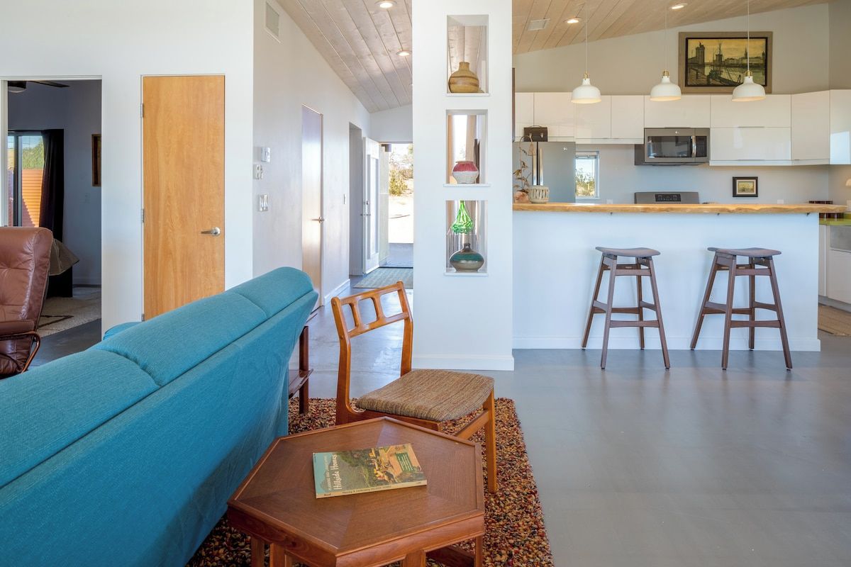A modern living room with a sofa, a wooden table, and a kitchen area with a bar stool and pendant lights.