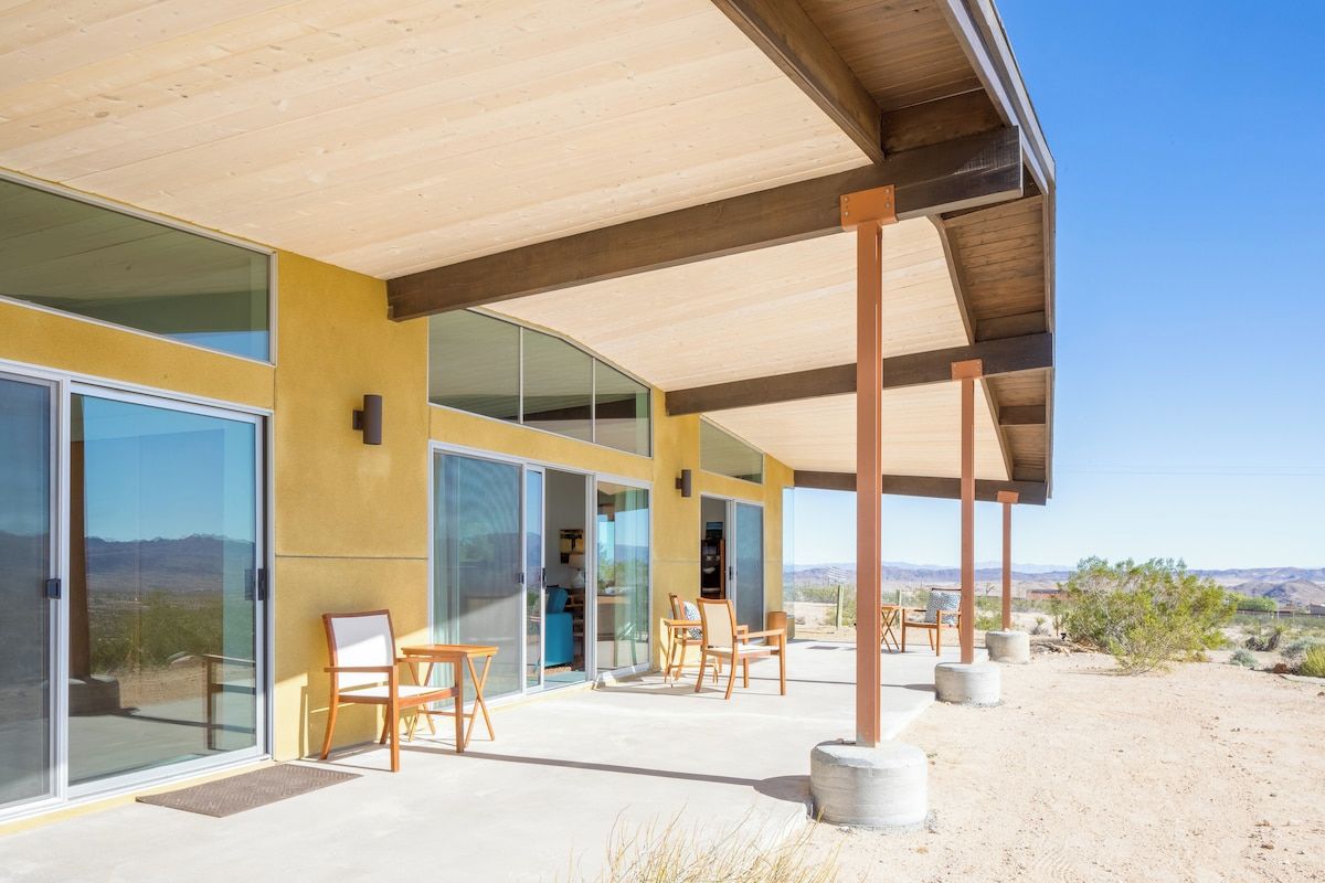 A modern building with large glass doors, a covered patio area with chairs, and a desert landscape in the background.