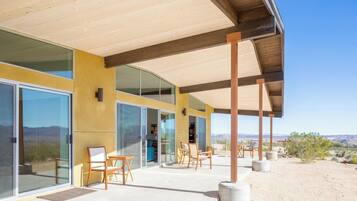 A modern building with large glass doors, a covered patio area with chairs, and a desert landscape in the background.