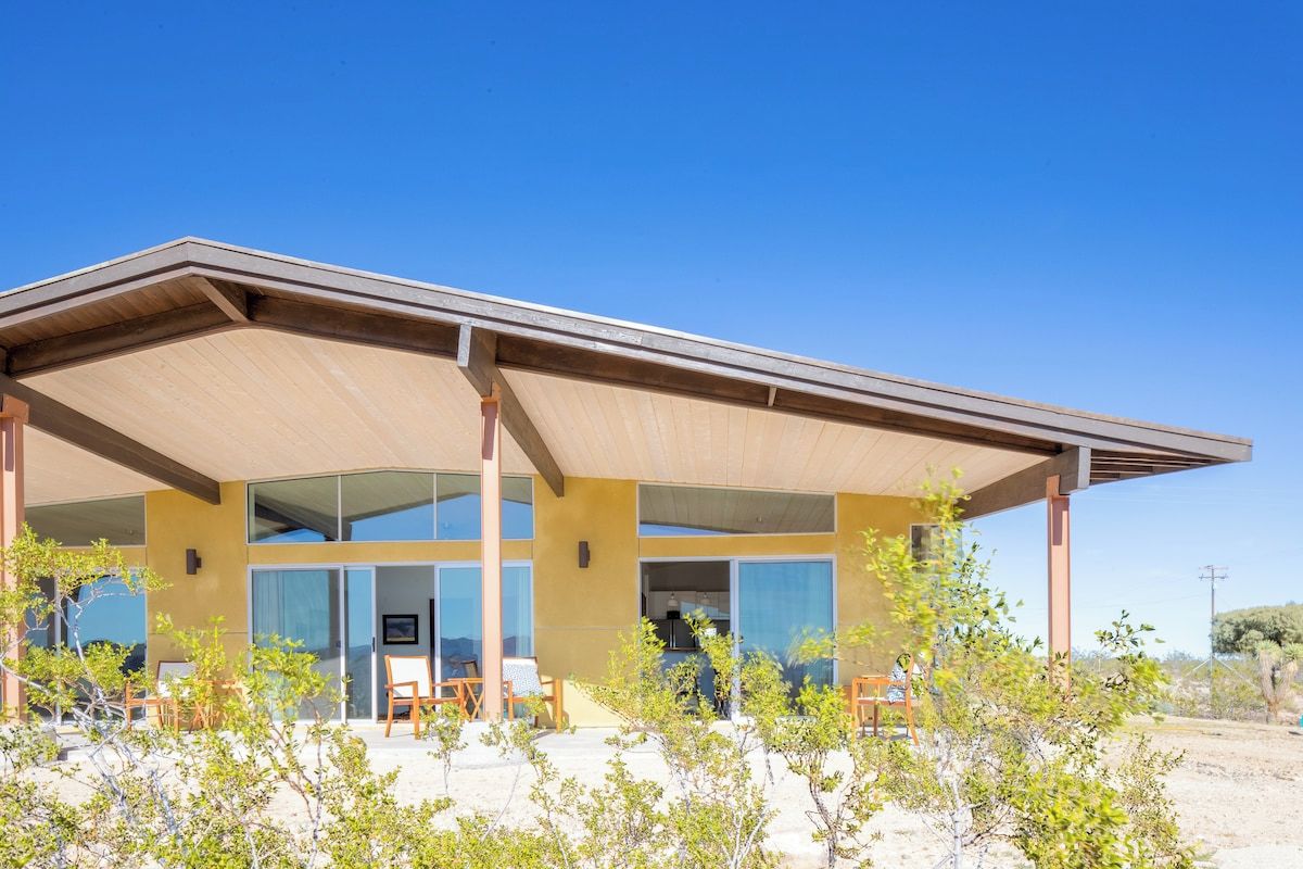 A modern building with a flat roof, large windows, and a covered patio area with chairs.