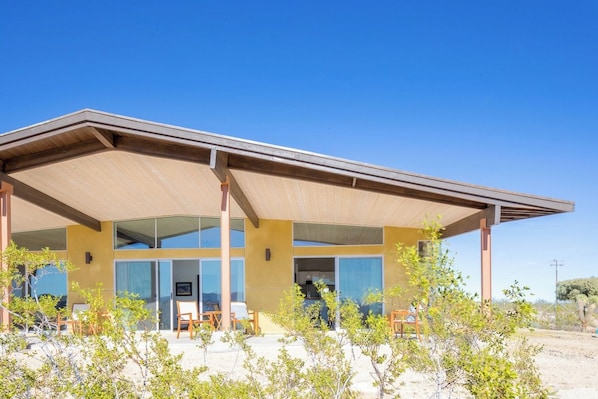 A modern building with a flat roof, large windows, and a covered patio area with chairs.