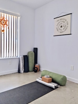 A room with a yoga mat, a green cushion, a black cushion, a kettlebell, and a wall-mounted light fixture.