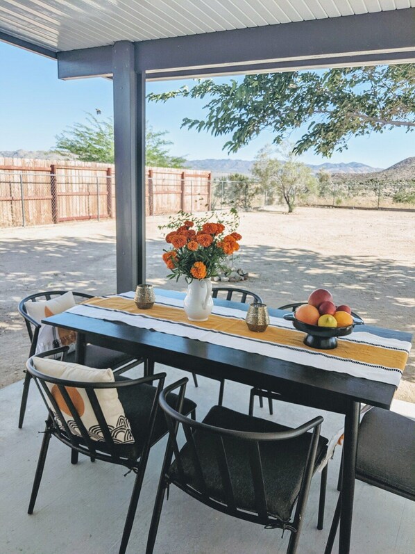 An outdoor patio with a table set for dining, chairs, a vase of flowers, and a fruit bowl.