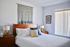A hotel room with a large bed, two bedside tables with lamps, a wall-mounted tapestry, and a window with blinds.