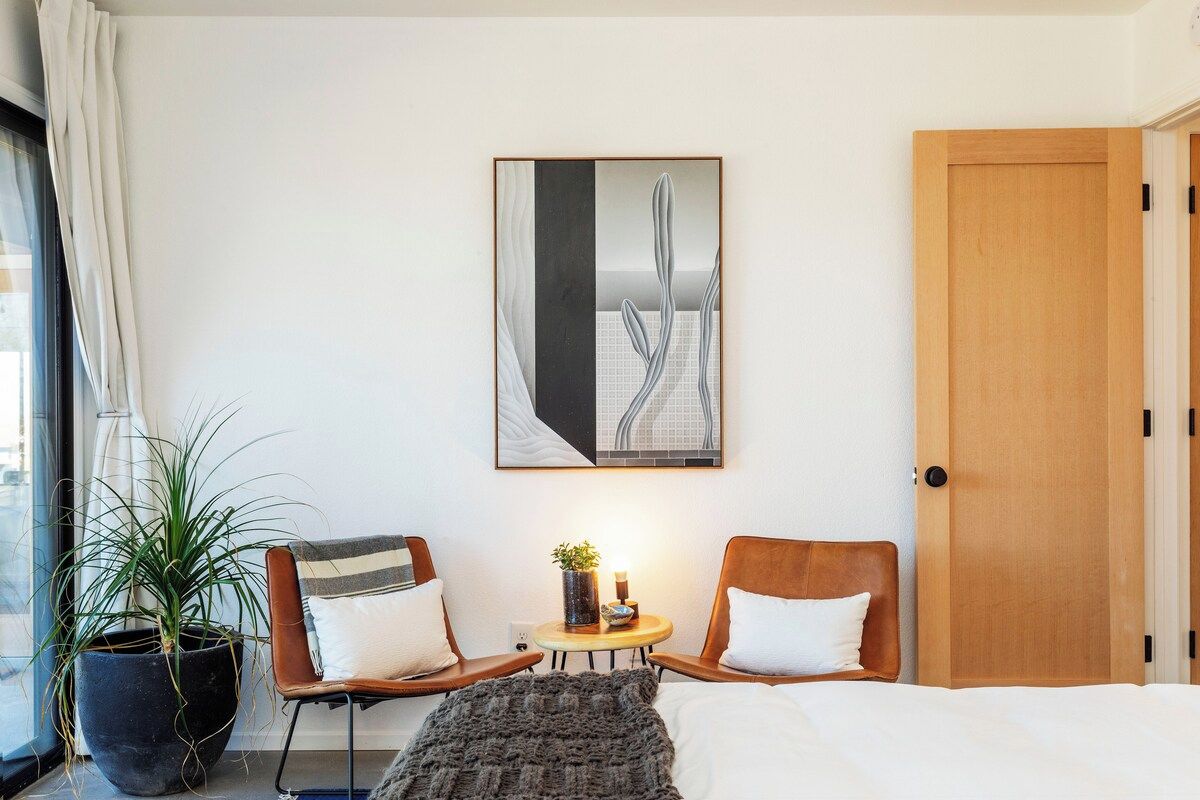 A hotel room with a bed, two chairs, a small table with a plant, a painting on the wall, and a large window with curtains.