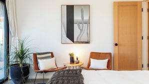 A hotel room with a bed, two chairs, a small table with a plant, a painting on the wall, and a large window with curtains.