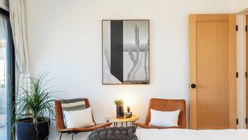 A hotel room with a bed, two chairs, a small table with a plant, a painting on the wall, and a large window with curtains.