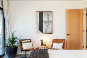A hotel room with a bed, two chairs, a small table with a plant, a painting on the wall, and a large window with curtains.