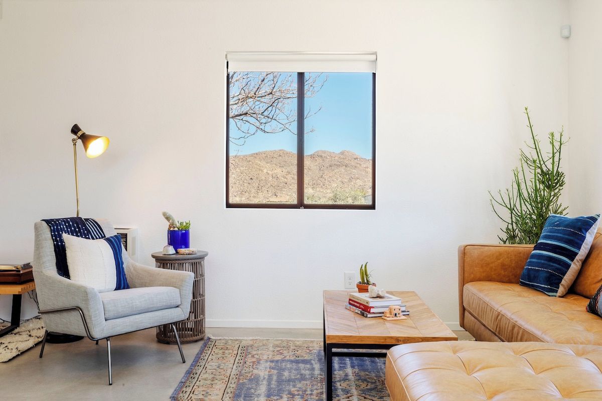 A living room with a sofa, armchair, coffee table, lamp, and a view of a desert landscape through a window.